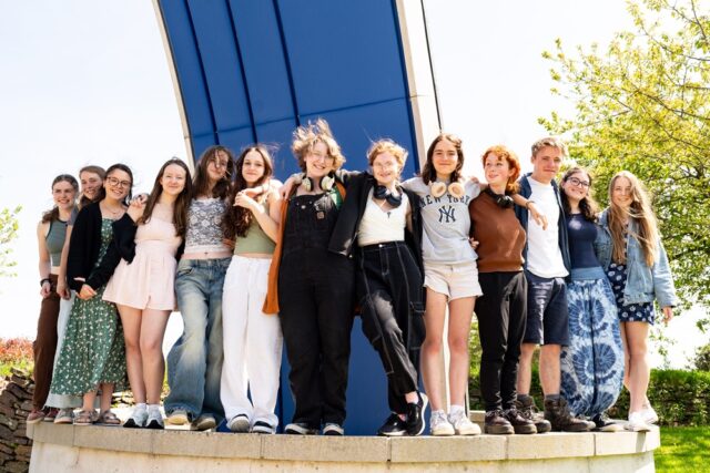 A group of students line up and stand on a low wall outside. They have their arms around each other.