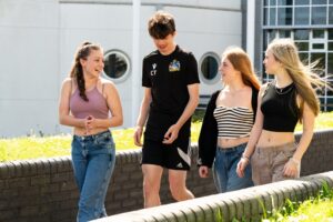 Four students walk outside in the sunshine. They all smile and laugh at each other as they walk.