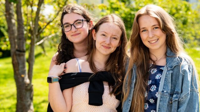 Three girls stand outside and face the camera. One girl stands behind another and puts her hands on her shoulders. They all smile.