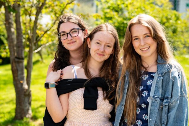 Three girls stand outside and face the camera. One girl stands behind another and puts her hands on her shoulders. They all smile.