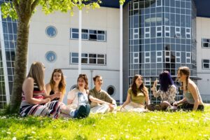 A group of students sit on the grass outside a college building. They have daisies surrounding them. They are all chatting and laughing.