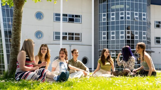 A group of students sit on the grass outside a college building. They have daisies surrounding them. They are all chatting and laughing.