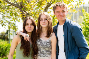 A group of three students stood outside in the sun. They all have their arms around each other and they smile at the camera.