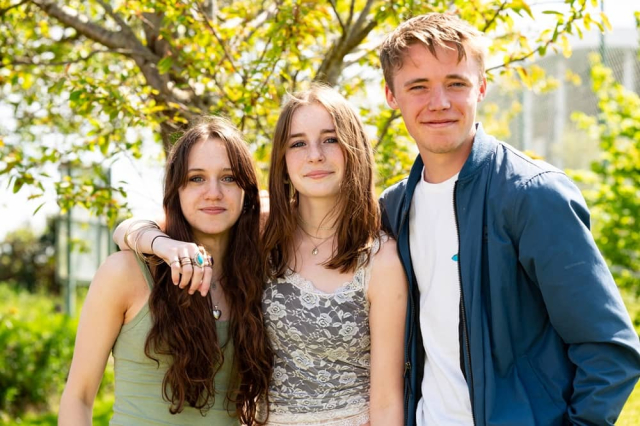 A group of three students stood outside in the sun. They all have their arms around each other and they smile at the camera.