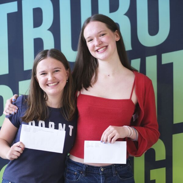 Two girls hold their results up to the camera. They are smiling and have their arms around each other.