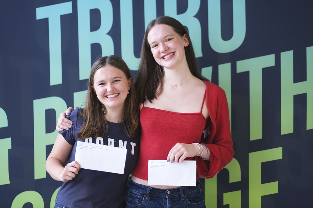 Two girls hold their results up to the camera. They are smiling and have their arms around each other.