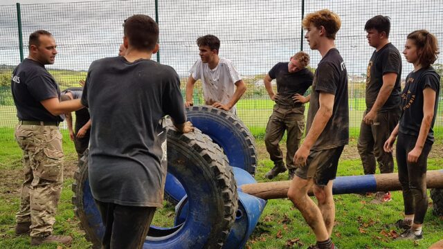 Students stood in a muddy field with large tyres and other sport equipment.