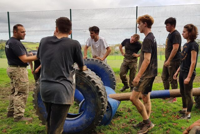 Students stood in a muddy field with large tyres and other sport equipment.