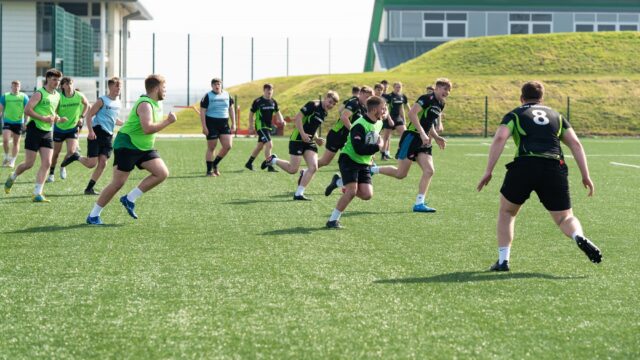 Rugby players are on the pitch dressed in green and black. They are on astroturf and the sky is cloudy.