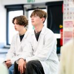 Two students in lab coats sit facing the front of the class.