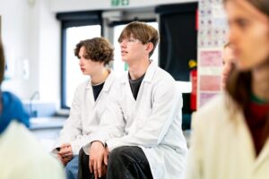 Two students in lab coats sit facing the front of the class.