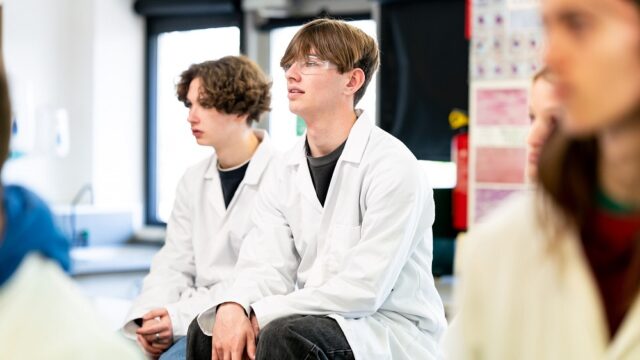 Two students in lab coats sit facing the front of the class.