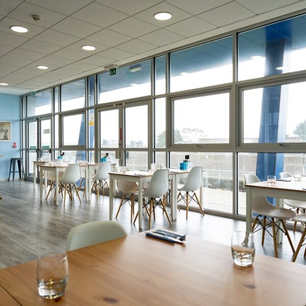 A photo of the inside of the Senara restaurant. There are wood tables and white chairs and large windows across the wall.