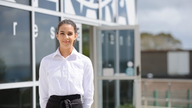 A photo of a female stood in front of the Senara restaurant. She is dressed in a white shirt and balck trousers with an apron tied around her waist. Her hands are behind her back.