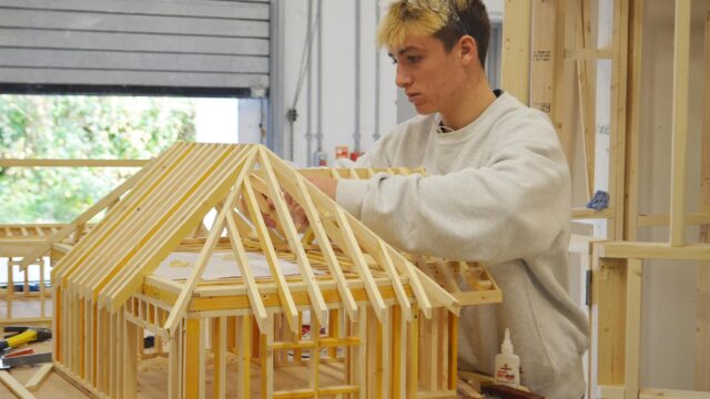 A student works on a small wooden structure of a building.