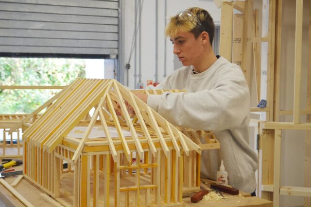 A student works on a small wooden structure of a building.