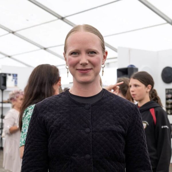 A student in a dark jacket stands in an art exhibition and smiles at the camera.
