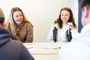 Two female students sit at a classroom table and smile at two students sat across from them, with their backs to the camera.