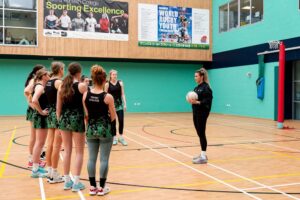 A team of female netball players stand in a semi circle facing another member of their team in a modern sports hall.