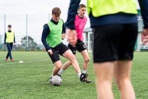 Male footballers wearing bright jerseys play on the AstroTurf at the college.