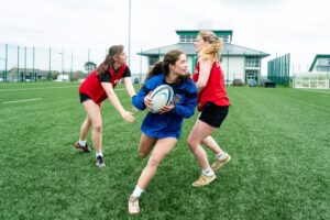 A female rugby player wearing a blue jersey dodges between two other female players wearing red jerseys on the AstroTurf.