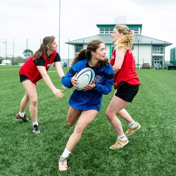 A female rugby player wearing a blue jersey dodges between two other female players wearing red jerseys on the AstroTurf.