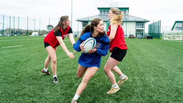 A female rugby player wearing a blue jersey dodges between two other female players wearing red jerseys on the AstroTurf.
