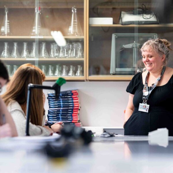 A tutor stands with her hands in her pocket looking at students working at a lab table. There are beakers in cabinets behind them.