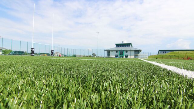 A close up of the grass on the astroturf with college buildings in the horizon.