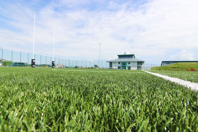 A close up of the grass on the astroturf with college buildings in the horizon.