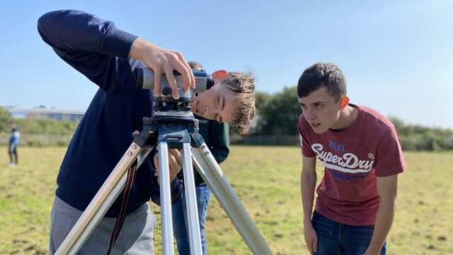 Students are fitting a device to a tripod in a large field.