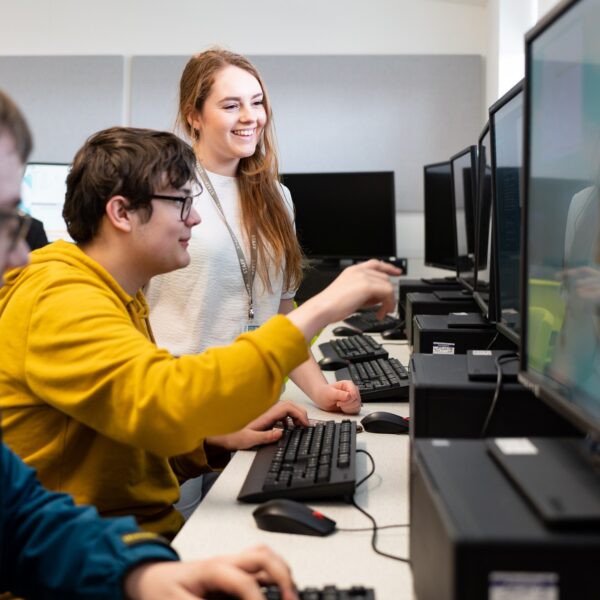 A tutor stands next to two students who are working at computers. One student points towards their computer screen.