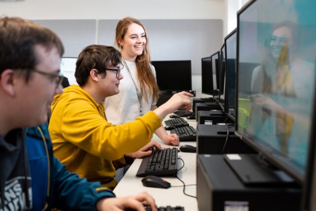 A tutor stands next to two students who are working at computers. One student points towards their computer screen.