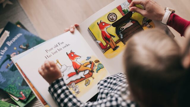 A child and an adult look at a picture book together. The page open shows a fox with the words 'Poor old fox has lost his socks'.