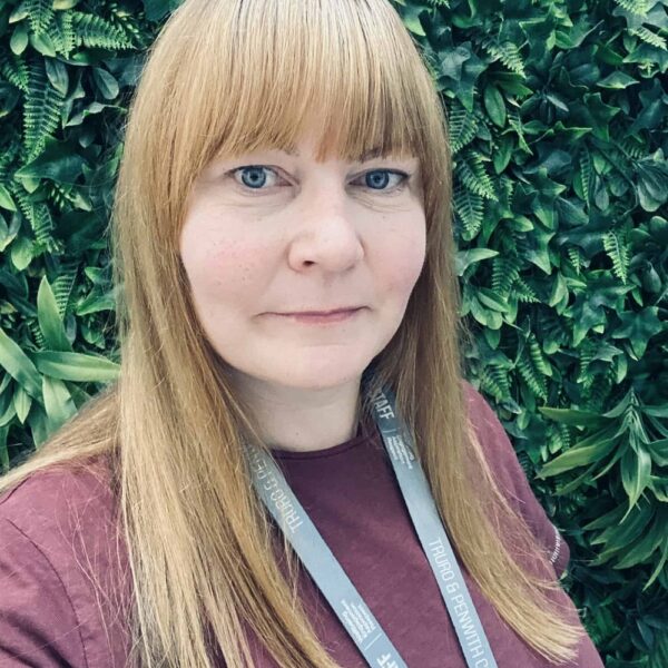 A photo of Tracey, she is wearing a red top and is stood in front of a green floral background.