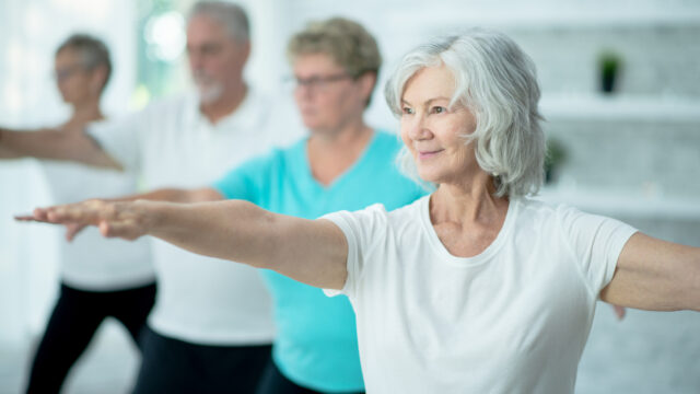 A small group of seniors stand evenly spready apart in a studio with their hands extended as they stretch and balance during a Tai Chi class. They are each dressed comfortably and are focused on the movements.