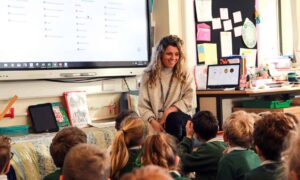 A teacher sits on a chair with primary school children sitting on the floor in front of her.