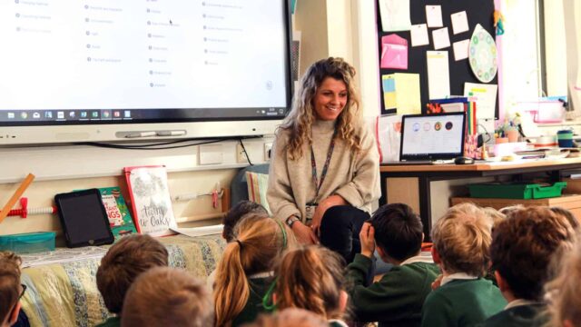 A teacher sits on a chair with primary school children sitting on the floor in front of her.