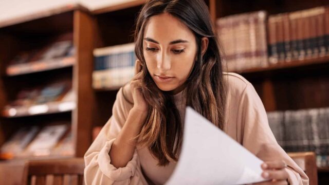 A young woman sits in a library and looks at a large book.