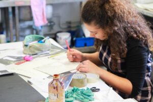 A student at an art table with pots of paint, tape, scissors, and paper.