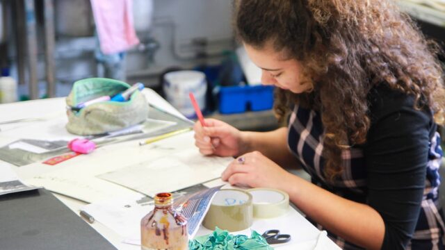 A student at an art table with pots of paint, tape, scissors, and paper.