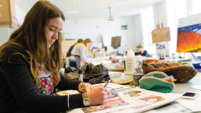 A student in an art class working in a sketchbook. There are other students at other tables.