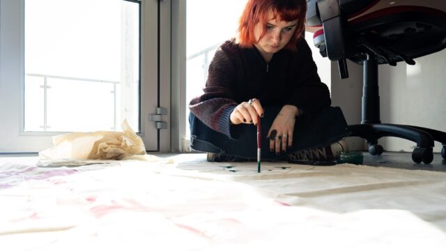 A student sits on the floor with a paintbrush in hand, painting on a large piece of material.
