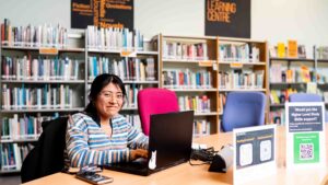 A higher education student sits at a table within a library with a laptop. She is wearing a jumper with stripes on it and is wearing glasses.