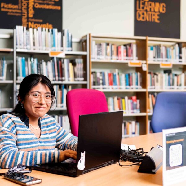 A higher education student sits at a table within a library with a laptop. She is wearing a jumper with stripes on it and is wearing glasses.