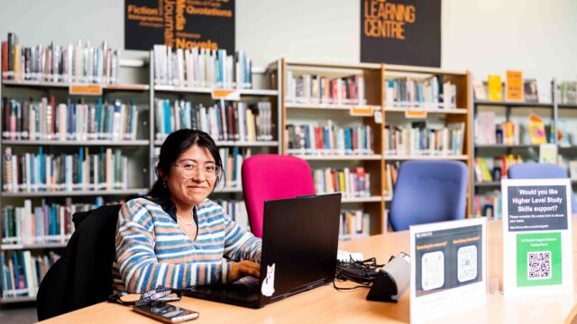 A higher education student sits at a table within a library with a laptop. She is wearing a jumper with stripes on it and is wearing glasses.