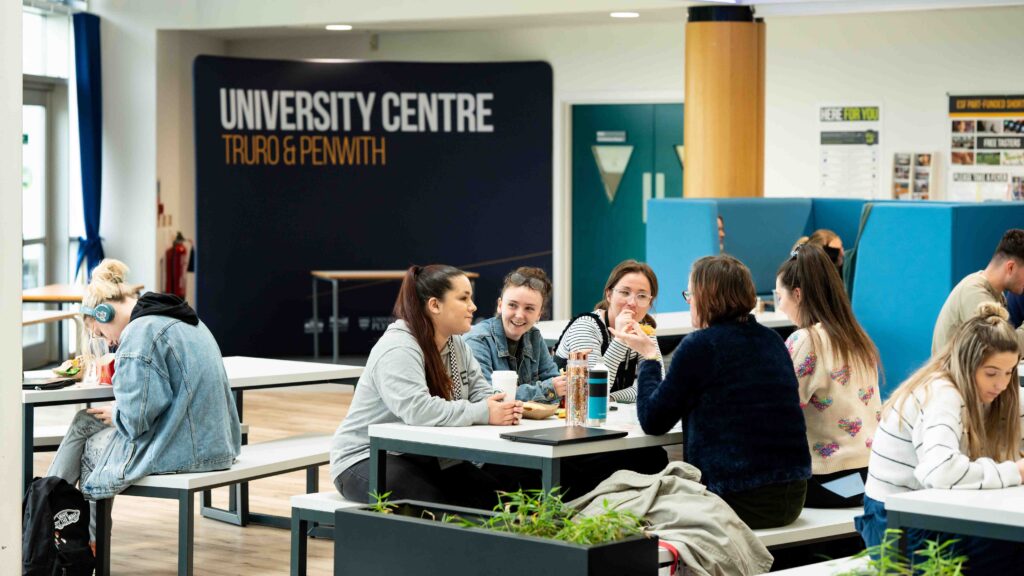 A group of students sit around large tables in the canteen area. There is a banner at the back of the room that says "University Centre Truro & Penwith".