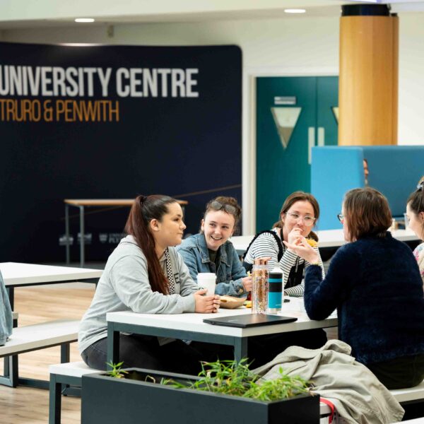 A group of students sit around large tables in the canteen area. There is a banner at the back of the room that says "University Centre Truro & Penwith".