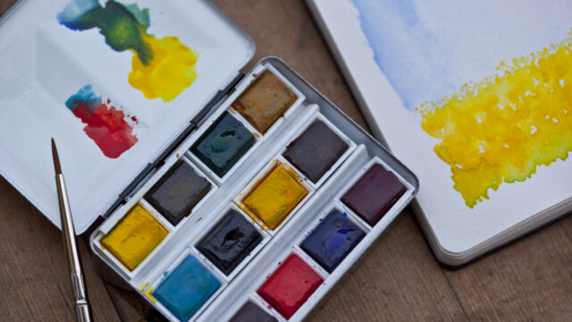 Watercolour paints in a palette, a sketchpad and a paintbrush.