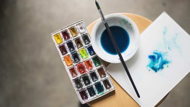 Artist's palette with painting colours, bowl with diluted blue paint and a paintbrush on a chair.